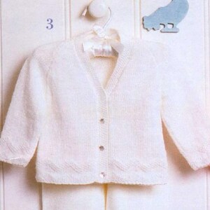 May include: A cream-colored, knitted baby cardigan with a V-neck and three small, clear buttons. The cardigan is hanging on a white hanger. The bottom edge has a scalloped design. The number "3" is visible in the upper left corner.