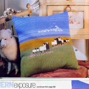 May include: A knitted pillow with a landscape design featuring sheep grazing in a field. The pillow has a blue sky, yellow and green fields, and white and black sheep. The pillow is resting on a red surface.