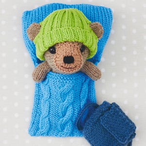May include: A knitted teddy bear wearing a green hat, nestled in a blue knitted sleeping bag with a cable knit design. A matching blue knitted bag is next to the sleeping bag. The bear has brown fur and a tan muzzle.