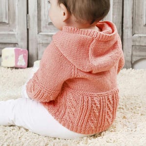 May include: A coral-colored knitted baby hoodie with a hood and intricate cable knit detailing on the lower back. The baby is wearing white pants and sitting on a white rug.
