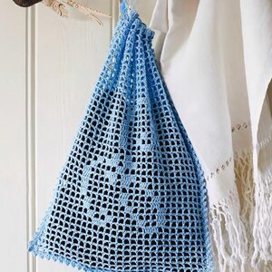 Op de afbeelding: Een lichtblauwe, open geweven gehaakte tas met een driehoekige vorm. De tas hangt aan een witte deur. De tas heeft een geschulpte rand en een decoratief patroon in het midden. Een witte handdoek hangt naast de tas.