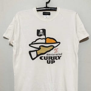 May include: White t-shirt featuring a graphic of a plate of food held by a hand, with a skull and crossbones flag. The text "mastermind CURRY UP" is below the graphic. The shirt has a crew neck and short sleeves.