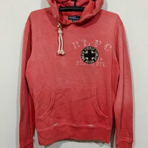 Könnte beinhalten: Roter Kapuzenpullover mit Fronttasche und Kordelzug. Der Pullover hat eine Grafik auf der Brust mit dem Text "RLPC LIFE GUARD BEACH DIV." und einem Kreuzemblem. Das Polo Ralph Lauren-Logo ist auf der Kapuze sichtbar.