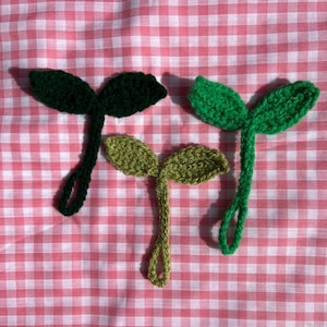 Op de afbeelding: Drie gehaakte plant- of scheutvormen met groene bladeren. De bladeren zijn gemaakt van garen en zijn bevestigd aan een stengel. De stengels zijn in verschillende kleuren: donkergroen, lichtgroen en lichtbruin.