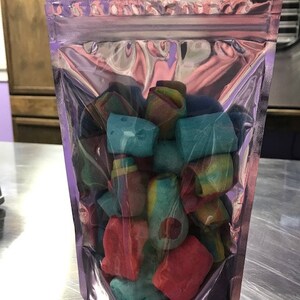 May include: A clear plastic resealable bag filled with colorful candy. The candy is in the shape of small, irregular blocks and is a mix of red, blue, green, and yellow.