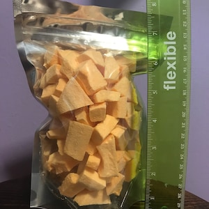 May include: A clear plastic bag filled with orange, cubed, dried fruit. The bag is standing upright next to a green ruler that measures 30 inches.