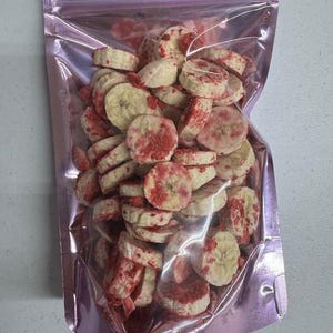 May include: A clear plastic bag filled with sliced, dried bananas with red flecks. The bag is sealed and has a pink metallic sheen. The banana slices are light yellow with red accents, suggesting a strawberry coating.