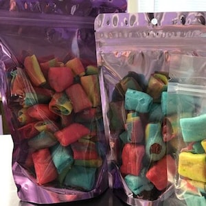 May include: Two clear plastic bags filled with colorful candy. The candy is shaped like small, rectangular pillows and is a mix of red, blue, green, and yellow.