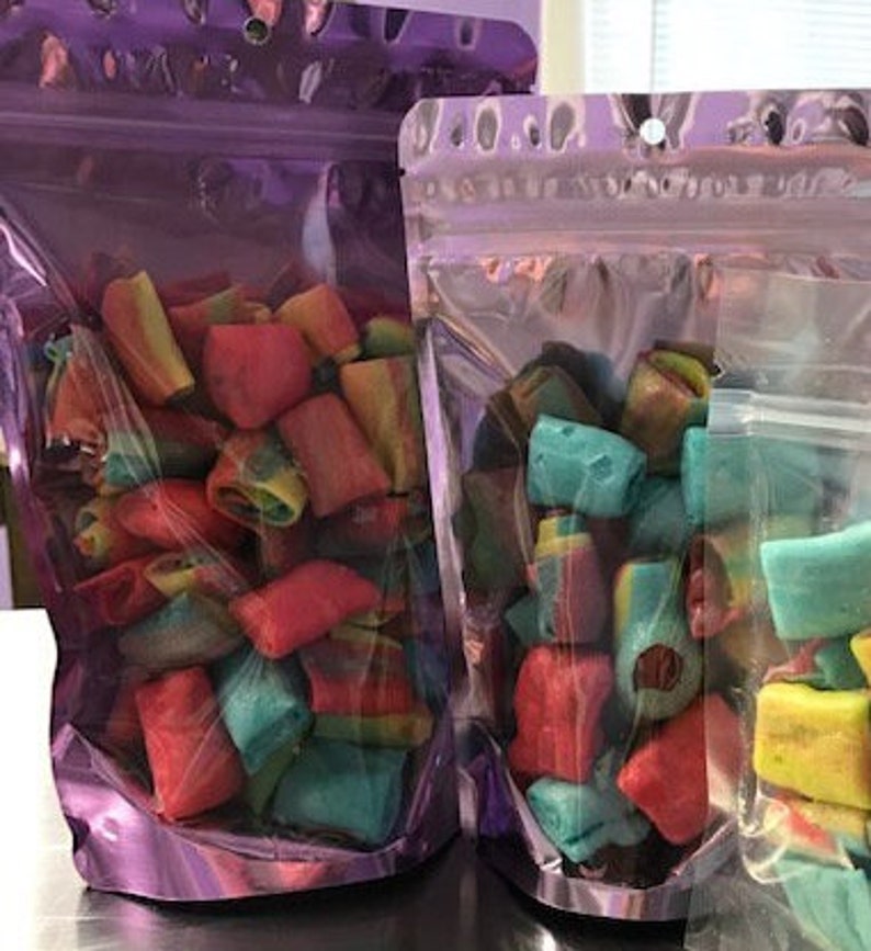 May include: Two clear plastic bags filled with colorful candy. The candy is shaped like small, rectangular pillows and is a mix of red, blue, green, and yellow.