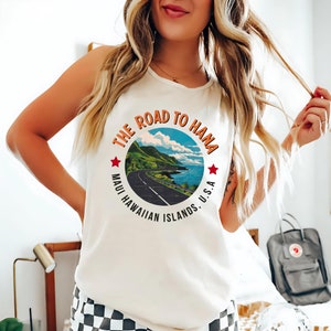 May include: White tank top with a graphic print of a road winding through a lush green landscape with the text "The Road to Hana, Maui Hawaiian Islands, U.S.A."