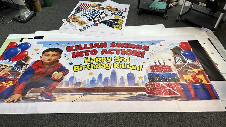 Puede incluir: Una pancarta de cumplea&ntilde;os rectangular de vinilo con un ni&ntilde;o en un disfraz de superh&eacute;roe rojo y azul. El texto dice "KILLIAN SWINGS INTO ACTION!" y "Happy 3rd Birthday Killian!" con un horizonte urbano, un pastel y regalos.