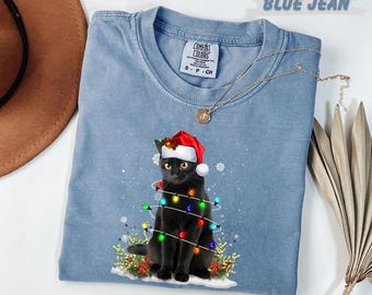 May include: A light blue, crew neck t-shirt with a graphic of a black cat wearing a Santa hat and wrapped in Christmas lights. The shirt has a tag that says "Comfort Colors". A gold necklace is draped across the shirt.