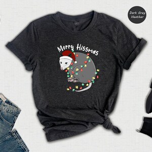 May include: A dark grey heather t-shirt with a white possum wearing a Santa hat and surrounded by Christmas lights. The text on the shirt reads 'Merry Hissmas'.