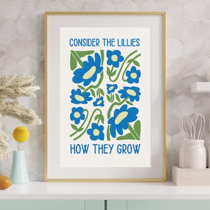 May include: A framed art print with a gold border features blue flowers and green stems on a white background. The text "CONSIDER THE LILLIES" and "HOW THEY GROW" are in blue. The artwork is displayed on a white shelf.