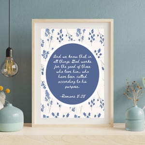 May include: Framed art print featuring a quote from Romans 8:28. The text is in a blue circle, surrounded by a floral pattern in blue on a cream background. The frame is a light wood color.