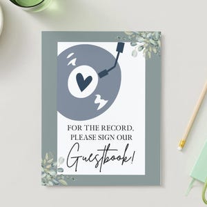 Puede incluir: Un letrero enmarcado con un borde gris y un interior blanco. El letrero presenta un diseño de disco azul con un corazón y el texto "FOR THE RECORD, PLEASE SIGN OUR Guestbook!" Acentos florales decorativos en las esquinas.