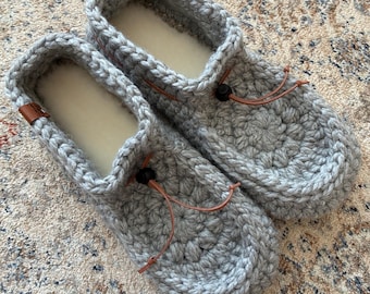 May include: A pair of light grey crochet slippers featuring a thick, textured yarn construction with a rounded toe design. Each slipper includes a brown leather cord drawstring with a black plastic toggle and a small brown leather brand tag.