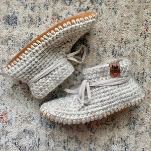 May include: A pair of light beige crochet ankle boots with a thick, textured knit pattern. Each boot features a matching beige drawstring tie and a tan leather-like patch on the side with a small tree graphic and the text "LUMBERJACKS CO."