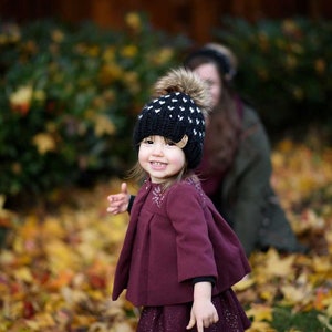 May include: A young girl wearing a black and white knitted hat with a white pom pom, a burgundy coat, and a sparkly burgundy skirt. She is standing in a pile of autumn leaves.