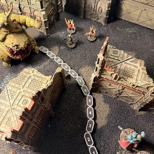 May include: A tabletop wargaming scene featuring a black movement gauge with white numbers 1 through 12, surrounded by miniature figures including a large yellow creature, several smaller warriors, and ruined building structures.
