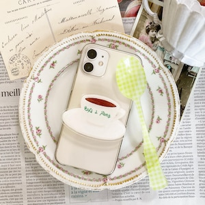 Puede incluir: Una funda de teléfono blanca con la parte posterior transparente y una pegatina de una taza de café con el texto "Café à Paris". La funda del teléfono está sobre un plato blanco con un patrón floral rosa y verde. Una cuchara de cuadros verdes y blancos está al lado de la funda del teléfono.