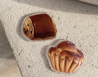 Puede incluir: Dos pegatinas de resina con ilustraciones de productos de panadería. Una pegatina muestra un rollo de pan con espiral de chocolate, la otra muestra un bollo redondo de brioche de color marrón dorado.