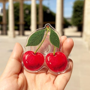 Peut inclure: Un support de téléphone en acrylique transparent présentant une illustration vive de deux cerises rouge vif avec des tiges et des feuilles vertes. Le design suit le contour du fruit, offrant une finition brillante sur le motif.
