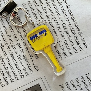 Peut inclure: Un porte-clés en forme de clé jaune et bleu avec le texte "LA POSTE". Le porte-clés est en acrylique transparent et possède un fermoir en métal argenté. Le design ressemble à une boîte aux lettres.