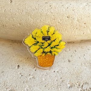 May include: A clear acrylic pin featuring a yellow mimosa flower arrangement in a woven basket. The pin has a small black label that reads "Mimosa 17€". The mimosa flowers are bright yellow, and the basket is a warm orange color.