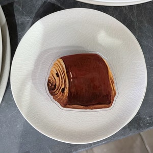 Puede incluir: Un plato de cerámica blanco con un pastel de chocolate marrón y blanco en la parte superior. El pastel tiene un diseño de espiral y está en un plato blanco con una superficie texturizada.