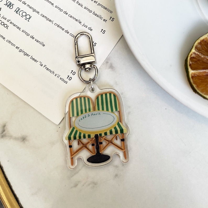 Op de afbeelding: Een transparante acryl sleutelhanger met een ontwerp van een groene en witte gestreepte caf&eacute;stoel en tafel. De sleutelhanger heeft de tekst "caf&eacute; &agrave; Paris" op de tafel.