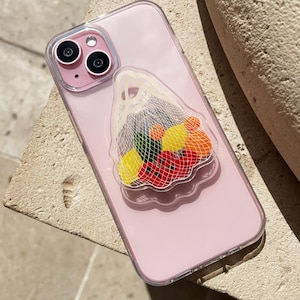 Puede incluir: Un teléfono inteligente rosa con una funda transparente. Una superposición transparente presenta una bolsa de malla llena de frutas coloridas, incluyendo pimientos rojos, naranjas y limones amarillos. El teléfono descansa sobre una superficie texturizada de color claro.