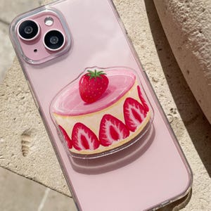 May include: Pink iPhone case with a clear, attached pop-socket featuring a strawberry cake design. The cake has a pink frosting layer, a whole strawberry on top, and strawberry slices around the sides.