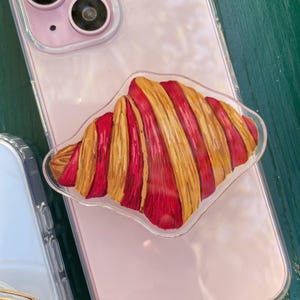 May include: A clear phone grip featuring a colorful illustration of a croissant. The grip is attached to a pink phone with a clear case. The croissant design has red and yellow stripes.