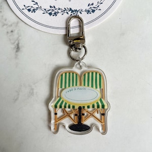 Op de afbeelding: Een transparante acryl sleutelhanger met een cartoonillustratie van een caf&eacute;tafel en twee stoelen met groen-wit gestreept stof. De tekst "caf&eacute; &agrave; Paris" is op de tafel gedrukt.