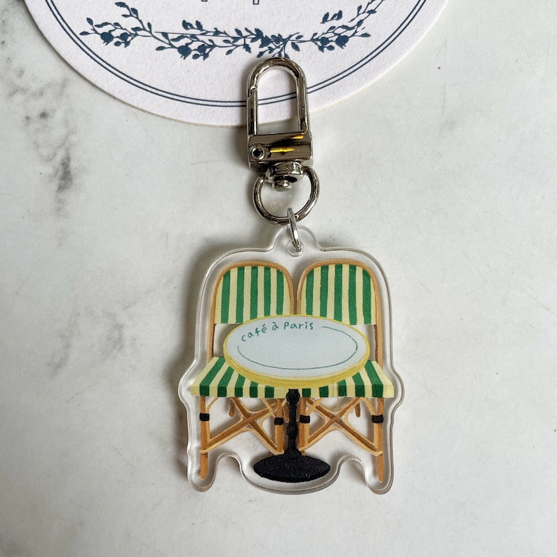 Op de afbeelding: Een transparante acryl sleutelhanger met een cartoonillustratie van een caf&eacute;tafel en twee stoelen met groen-wit gestreept stof. De tekst "caf&eacute; &agrave; Paris" is op de tafel gedrukt.