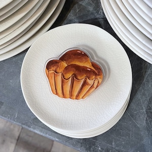 May include: A stack of white ceramic plates with a textured, crackled surface. A clear sticker of a golden-brown pastry is on top of one plate. The pastry has a detailed, painted appearance, resembling a brioche or similar baked good.