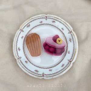 May include: A white porcelain plate with a pink floral pattern features two decorative food items. One is a brown madeleine cookie and the other is a pink and red raspberry cake with a heart-shaped detail.