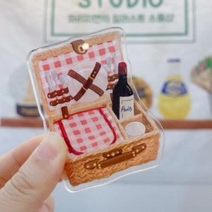 May include: A brown wicker picnic basket with a red and white checkered lining. The basket is open and contains a plate, silverware, a bottle of wine labeled "Paris", and a small white container.