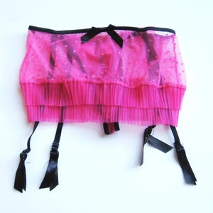 May include: A bright pink garter belt with black accents. The belt features a layered, pleated design with a sheer overlay dotted with small white spots. Black satin ribbons and adjustable straps complete the design.