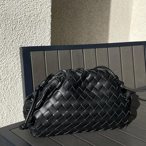 May include: Black woven leather clutch bag with a drawstring closure. The bag is rectangular with a rounded top and is resting on a dark wooden surface. The woven pattern creates a textured appearance.