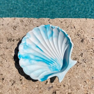 Op de afbeelding: Een witte schelpvormige schaal met blauwe marmering. De schaal is gemaakt van keramiek en heeft een gladde, glanzende afwerking.