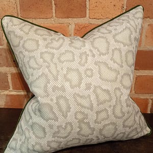 May include: A decorative square pillow with a light gray animal print pattern. The pillow has a dark green trim and is set against a brick wall background. The pillow is a home decor item.