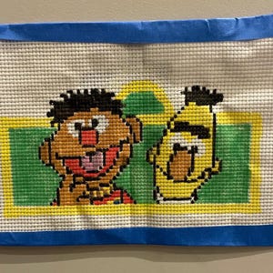 May include: Handmade needlepoint featuring two cartoon characters, one with brown skin and black hair, the other yellow with black hair. The characters are set against a green and yellow background, framed by a blue border.