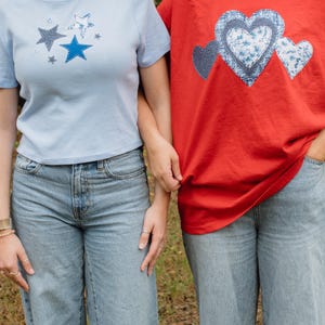 Könnte beinhalten: Zwei Personen tragen hellblaue Jeans. Eine Person trägt ein hellblaues T-Shirt mit Sterndesigns. Die andere Person trägt ein rotes T-Shirt mit Herzdesigns. Beide Shirts sind kurzärmelig.
