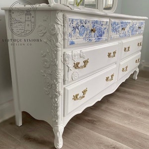 May include: White dresser with ornate floral carvings and gold hardware. The top drawers have a blue and white floral pattern. The dresser is labelled "Vintage Visions Design Co."