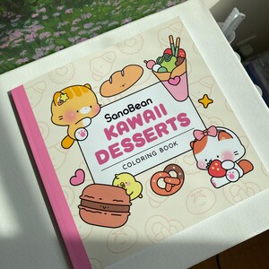 K&ouml;nnte beinhalten: Ein "SanoBean Kawaii Desserts Malbuch" mit rosa Buchr&uuml;cken. Das Cover zeigt Cartoon-Illustrationen von Desserts, darunter eine Brezel, ein Macaron und Eis. Zwei Cartoon-Katzen und ein K&uuml;ken sind ebenfalls abgebildet.