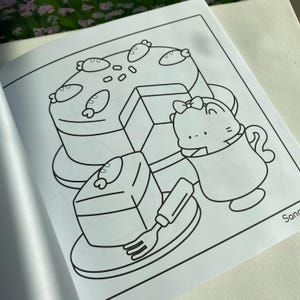 K&ouml;nnte beinhalten: Schwarz-Wei&szlig;-Illustration eines Kuchens, eines Kuchenst&uuml;cks und einer Figur in einer Teetasse. Der Kuchen ist mit fischf&ouml;rmigen Designs verziert. Eine Gabel liegt auf einem Teller neben dem Kuchenst&uuml;ck. Die Figur hat eine Schleife.
