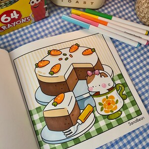 K&ouml;nnte beinhalten: Eine Malbuchseite mit einem Cartoon-Karottenkuchen mit Scheiben, einer Teetasse mit einer Katze und einer Gabel. Die Seite befindet sich auf einer blau-wei&szlig; karierten Oberfl&auml;che, mit Markern und Buntstiften in der N&auml;he.