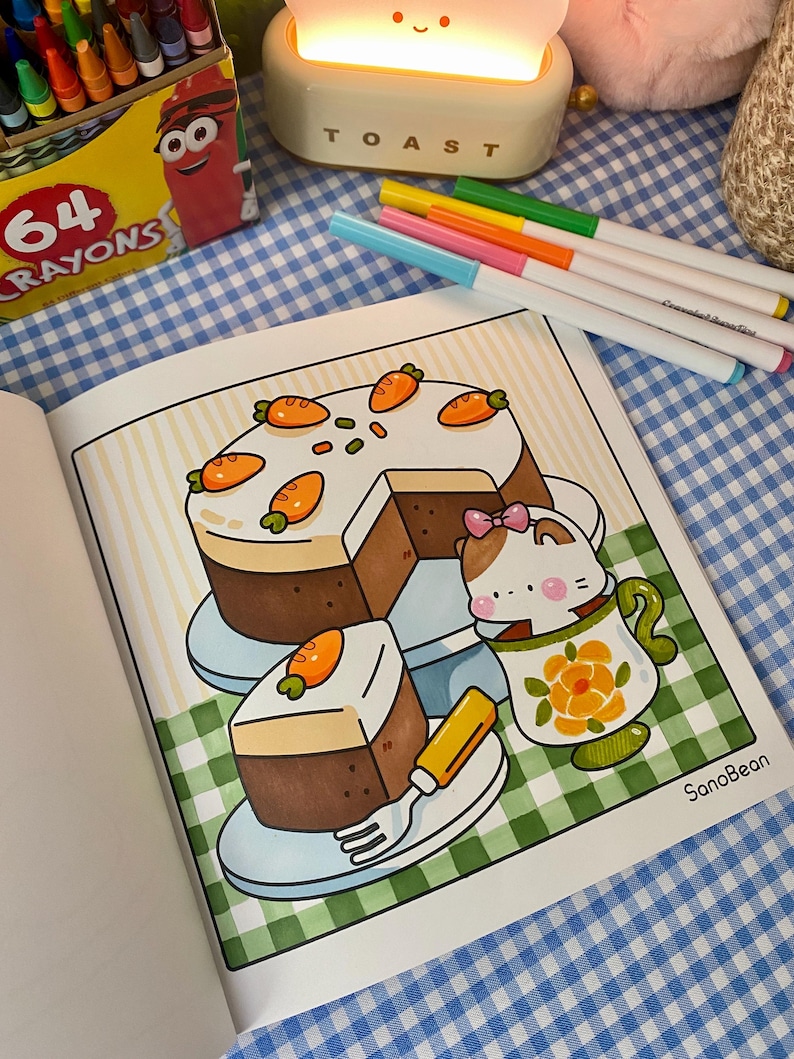 K&ouml;nnte beinhalten: Eine Malbuchseite mit einem Cartoon-Karottenkuchen mit Scheiben, einer Teetasse mit einer Katze und einer Gabel. Die Seite befindet sich auf einer blau-wei&szlig; karierten Oberfl&auml;che, mit Markern und Buntstiften in der N&auml;he.